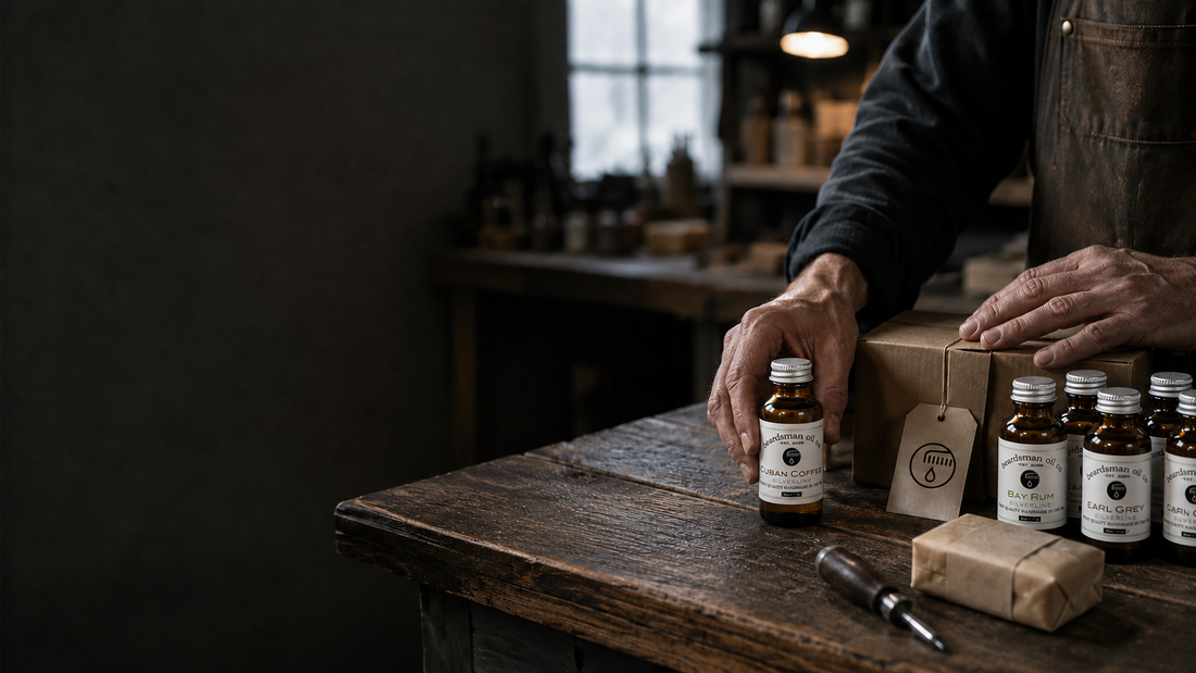 A craftsman’s weathered hands at a wooden workbench, picking up a bottle of Beardsman Cuban Coffee beard oil beside bottles of Bay Rum and Earl Grey, a hand-wrapped bar of soap, and twine-tied kraft boxes with the Beardsman drop icon.